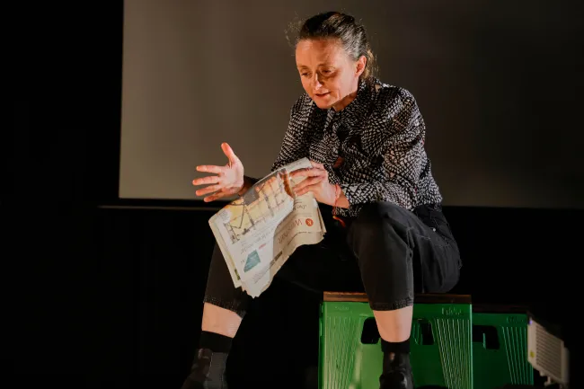 Schauspielerin Gerti Drassl sitzt auf einem Hocker auf der Bühne und hält eine Zeitung in der Hand, sie sieht Richtung Boden