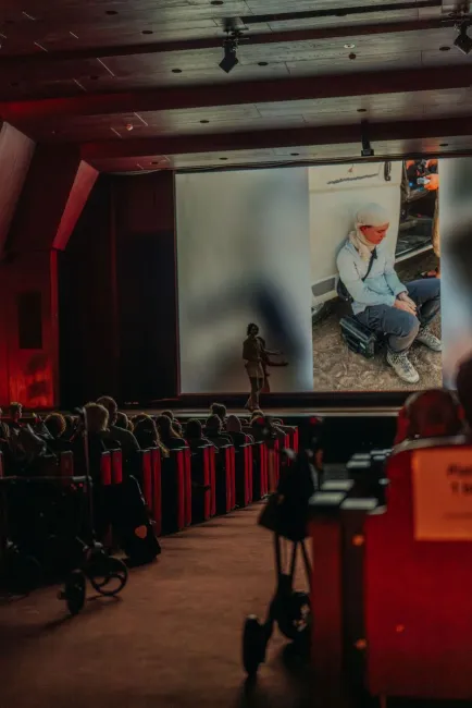 Der Publikumsbereich in einem Saal wird von der letzten Reihe aus gezeigt. Auf der Bühne steht Schneider, hinter ihr ist auf eine große Leinwand ein Bild von einer Person, die vor einem Auto auf einer Box sitzt, projiziert