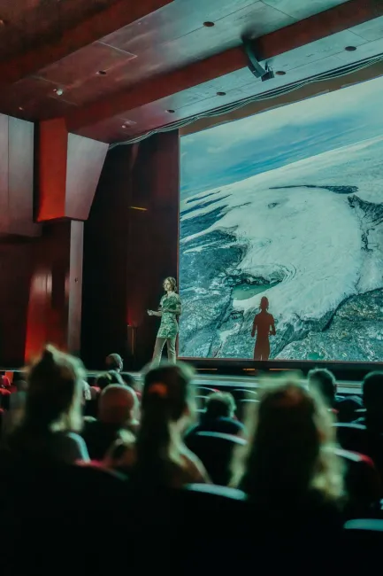 Louisa Schneider steht auf einer Bühne. Vor ihr sitzen Menschen im Publikum. Hinter ihr ist eine große Leinwand, auf der ein Bild einer schneebedeckten Landschaft gezeigt wird